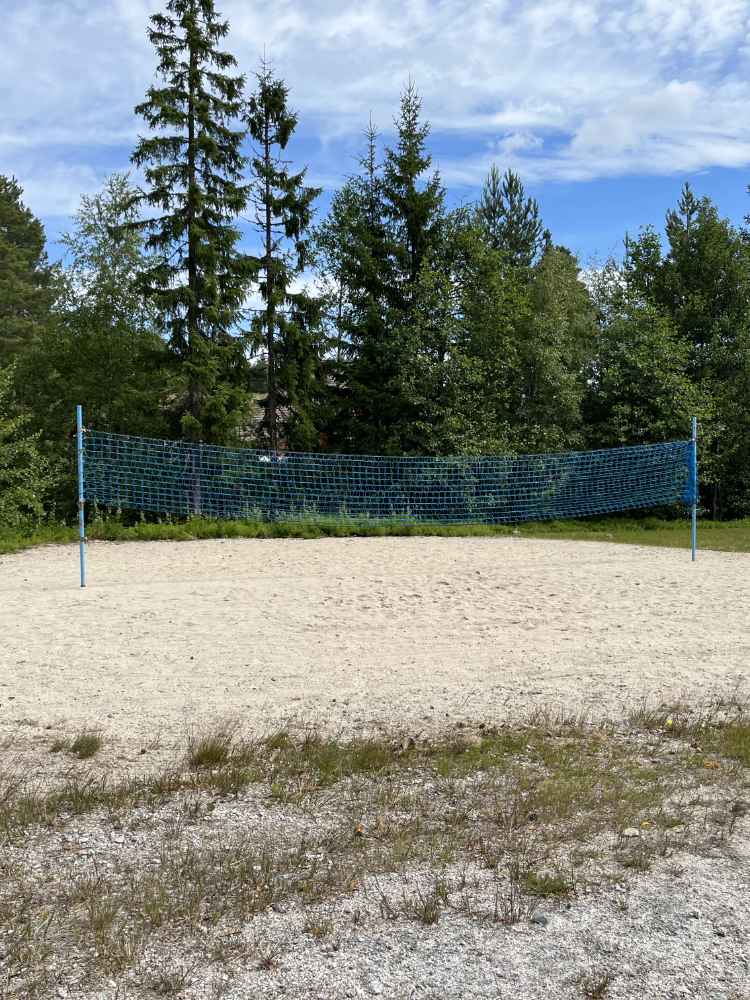 Blue volleyball net on sandy court surrounded by green grass, under a clear blue sky with a few clouds.