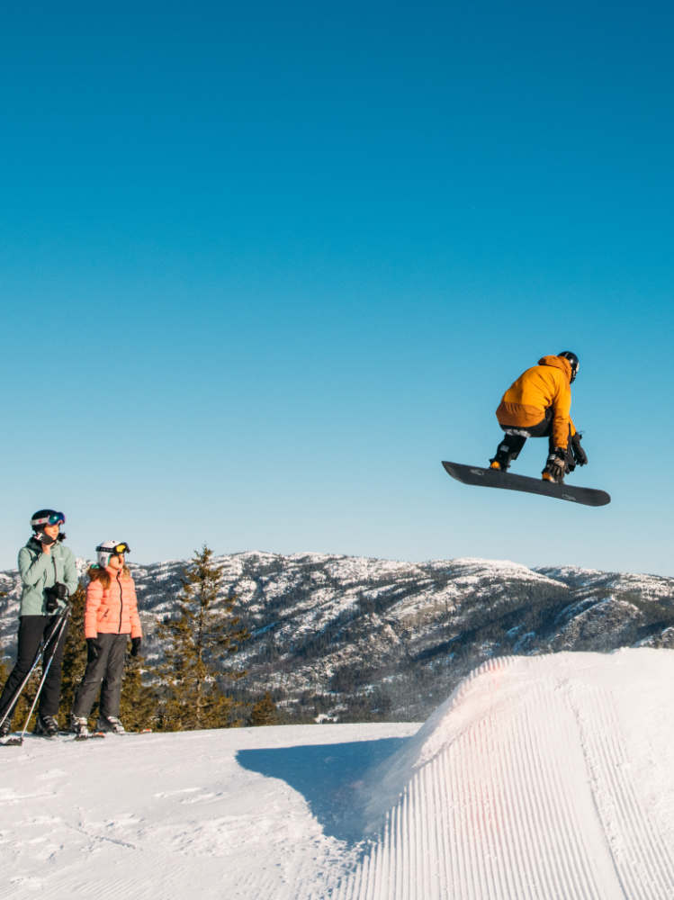 Snowboarder jumping over a ski ramp with friends watching under a blue sky